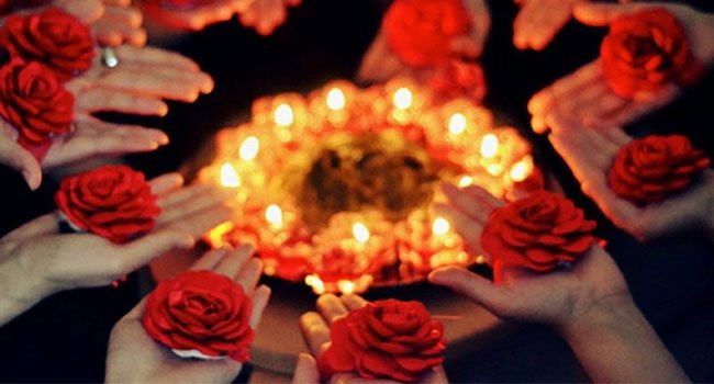 أيدٍ مزينة بالورود الحمراء حول صينية حناء مضيئة في ليلة الحناء.