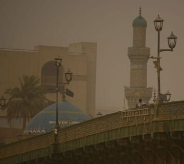 منظر لجسر الأحرار في بغداد يظهر تحت سماء مغبرة وضبابية مع مأذنة وقبة مسجد ملونة بالأزرق والذهبي في الخلفية 
