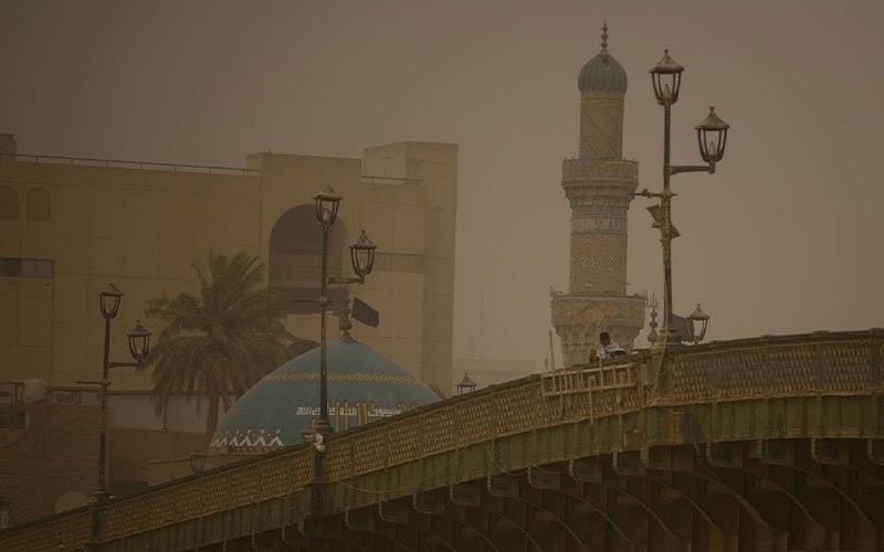 منظر لجسر الأحرار في بغداد يظهر تحت سماء مغبرة وضبابية مع مأذنة وقبة مسجد ملونة بالأزرق والذهبي في الخلفية 