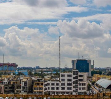 طقس-العراق منظر أفق مدينة عراقي تحت سماء زرقاء غائمة جزئياً تتخللها سحب بيضاء، مع مبانٍ سكنية وتجارية متفرقة وأشجار ونخل