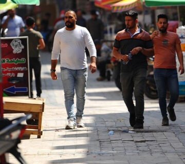 ثلاثة رجال يسيرون في شارع سوق شعبي مشمس ومظلل بالمظلات الملونة في إحدى المدن العراقية خلال فترة الظهيرة 