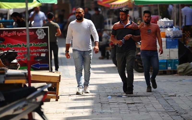 ثلاثة رجال يسيرون في شارع سوق شعبي مشمس ومظلل بالمظلات الملونة في إحدى المدن العراقية خلال فترة الظهيرة 