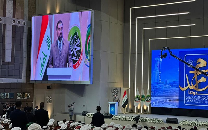 الحلبوسي يلقي كلمة في مؤتمر رسمي خلفه شاشة بعلم العراق 