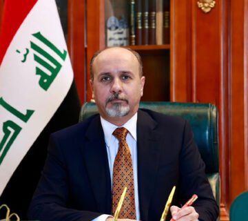 أمين بغداد يجلس في مكتبه وبجانبه العلم العراقي 