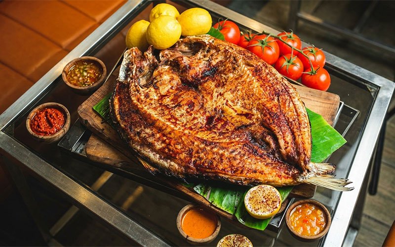 سمكة مسكوف عراقية مشوية محمرة على لوح خشبي، تحيط بها ثمار الليمون والطماطم الطازجة وأطباق صغيرة من الصلصات المتنوعة 