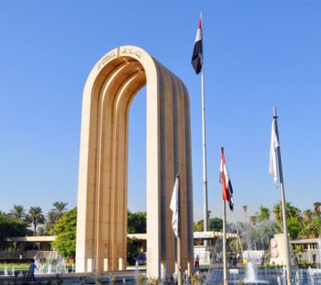 القوس المعماري والأعلام الوطنية عند مدخل جامعة بغداد تحت سماء صافية 