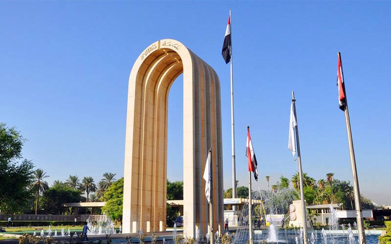 القوس المعماري والأعلام الوطنية عند مدخل جامعة بغداد تحت سماء صافية 