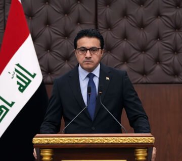 مسؤول عراقي (العبودي) يقف خلف منصة إلقاء كلمة وبجانبه علم العراق 