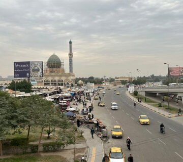 مشهد عام لشارع رئيسي وجسر في بغداد تحت سماء غائمة جزئياً، ويظهر جامع الشواف مع حركة السيارات والمشاة في المنطقة 