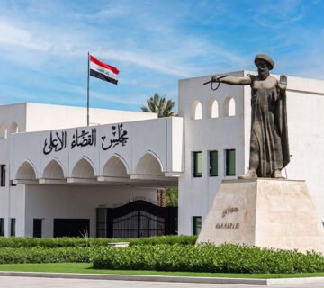 مبنى مجلس القضاء الأعلى في العراق مع علم العراق مرفوعاً أمامه، وتمثال الملك حمورابي واقفا على قاعدة حجرية في المقدمة 