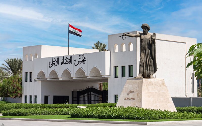 مبنى مجلس القضاء الأعلى في العراق مع علم العراق مرفوعاً أمامه، وتمثال الملك حمورابي واقفا على قاعدة حجرية في المقدمة 