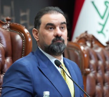 صورة لرئيس مجلس النواب العراقي السابق محمد الحلبوسي يرتدي بدلة زرقاء وربطة عنق صفراء، يجلس خلف الميكروفون وأمامه علم العراق 