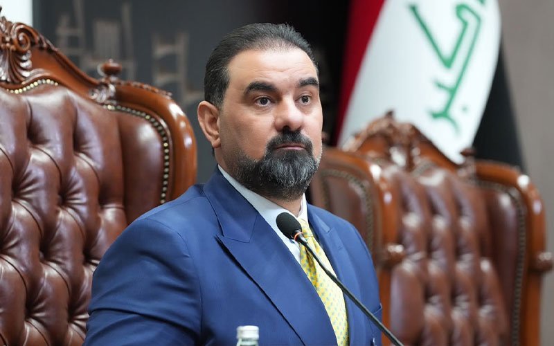 صورة لرئيس مجلس النواب العراقي السابق محمد الحلبوسي يرتدي بدلة زرقاء وربطة عنق صفراء، يجلس خلف الميكروفون وأمامه علم العراق 